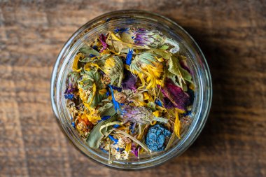 Dry flower and herbal tea leaves in a glass jar on wooden background, copy space. Herbal collection of chamomile, cornflower, mint, sea buckthorn, lemongrass, wild rose, dried citrus fruits and apple