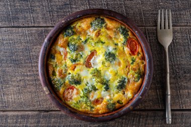 Ceramic bowl with vegetable frittata, simple vegetarian food. Frittata with egg, tomato, pepper, onion, green broccoli and cheese on wooden table, close up, top view. Italian egg omelette
