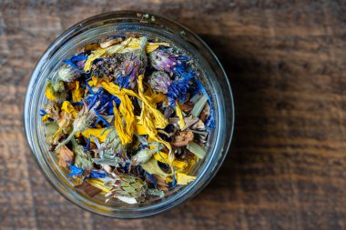 Dry flower and herbal tea leaves in a glass jar on wooden background, copy space. Herbal collection of chamomile, cornflower, mint, sea buckthorn, lemongrass, wild rose, dried citrus fruits and apple