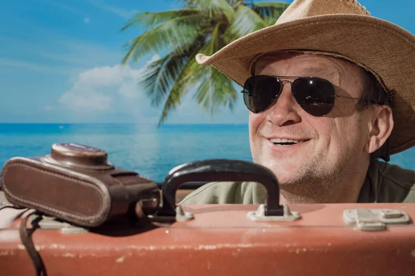 在热带海滩的阳光明媚的夏日 一个戴着太阳镜 头戴礼帽 头戴手提箱和照相相机的快乐老人出现在热带海滩 旁边是大海 棕榈树和蓝天的背景 度假和旅行概念 — 图库照片