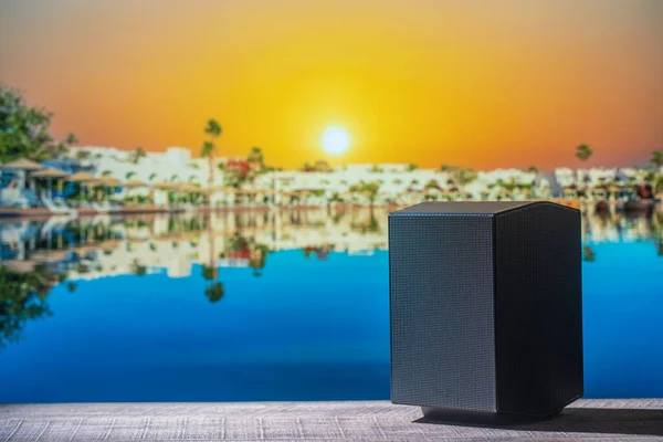 Black acoustic sound speaker on a wooden table against the tropical beach background with sea water during sunrise. Musical equipment, close up