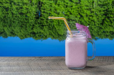 Güneşli bir yaz gününde, göl suyu ve orman arka planıyla ahşap masada cam bardakta duran sağlıklı, soğuk çilekli smoothie. Kapatın.
