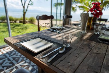 Boş bardaklar, beyaz tabaklar ve çatal bıçak Tayland 'ın tropik adasında güneşli bir yaz gününde bir plaj restoranında ahşap bir masada. Kapat.