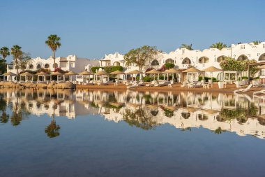 Gündoğumunda, Mısır 'ın Sharm El Sheikh tatil beldesindeki sakin deniz suyuna binalar, güneşlikler ve şemsiyeler yansıtılır.