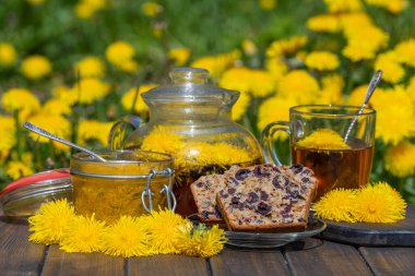 Sağlıklı karahindiba çiçeği çayı ahşap masada cam çaydanlıkta tatlı reçel ve vişneli çörekle birlikte bahar bahçesinde yakın.