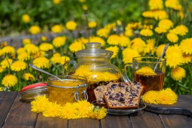 Sağlıklı karahindiba çiçeği çayı ahşap masada cam bir fincanda tatlı reçelli ve vişneli kek ile bahar bahçesinde, kapat