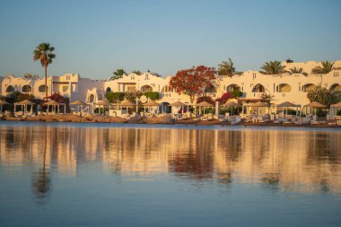 Gündoğumunda, Mısır 'ın Sharm El Sheikh tatil beldesindeki sakin deniz suyuna binalar, güneşlikler ve şemsiyeler yansıtılır.