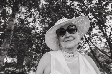 Doğanın arka planında hasır şapkalı, 65-70 yaşlarında mutlu yaşlı bir kadının portresi. Yaklaş. Siyah ve beyaz