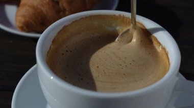 Tahta masada beyaz bardakta kaşıkla karıştırılmış koyu köpüklü sıcak kahve kapuçinosu, yakın çekim. Süt köpüğü kapuçino, latte.