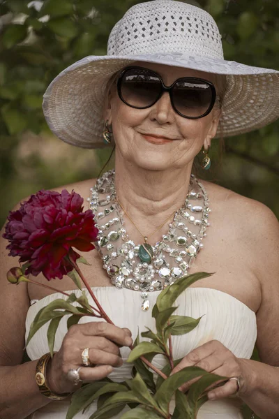 Portrait of a happy elderly woman 65 - 70 years old in a white straw hat with red peony flower, close up