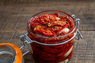 Sarımsaklı, zeytinyağlı ve ahşap bir masada cam kavanozda baharatlı kurutulmuş kırmızı domatesler. Kırsal stil, yakın çekim