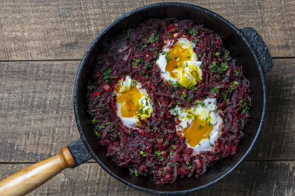 Boiled red beets with fried eggs, red peppers, onions and green dill in a frying pan on a wooden table, close up, top view. Food background. Healthy food