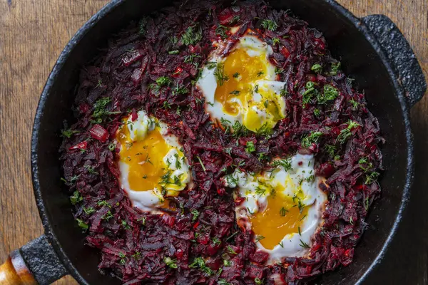 Boiled red beets with fried eggs, red peppers, onions and green dill in a frying pan on a wooden table, close up, top view. Food background. Healthy food