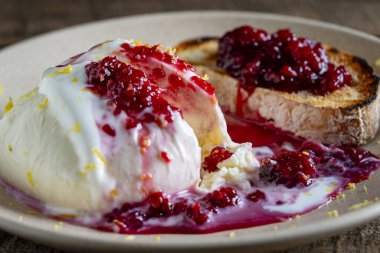 Beyaz mozzarella peyniri, ahududu reçeli ve tabakta bir dilim tam tahıllı ekmek ile servis edilir.