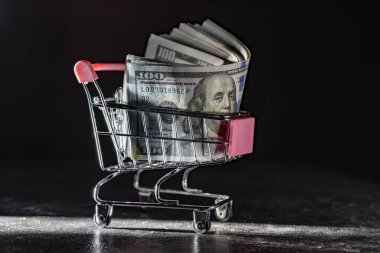 Tekerlekli alışveriş arabasının arka planında dolar banknotları var. Kapatın, kopyalayın. Finans, iş, alım-satım, harcama ve satış kavramı