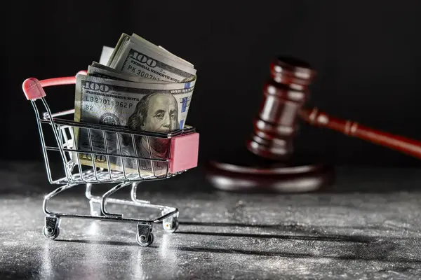 Shopping cart on wheels with dollar bills and wooden judge gavel on dark background, close up. Finance, business, purchases, spending and sales concept