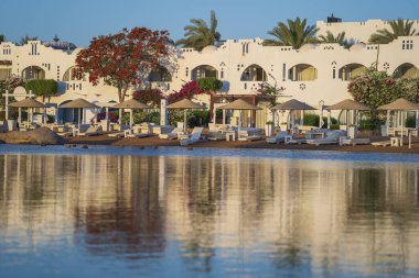 Gündoğumunda, Mısır 'ın Sharm El Sheikh tatil beldesindeki sakin deniz suyuna binalar, güneşlikler ve şemsiyeler yansıtılır.