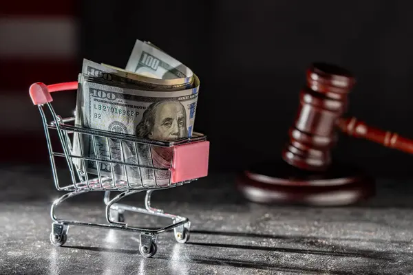 Shopping cart on wheels with dollar bills and wooden judge gavel on dark background, close up. Finance, business, purchases, spending and sales concept