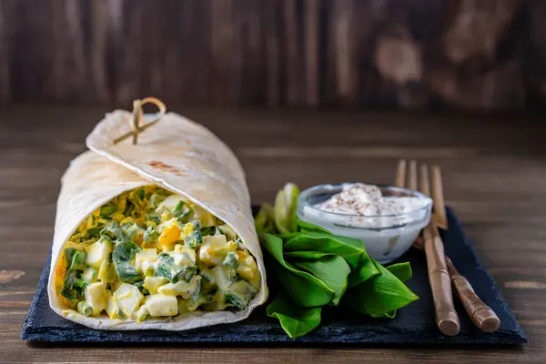 用煮熟的鸡蛋 绿色大蒜和酸奶油包裹自制卷饼 在木板上享用健康早餐 图库图片