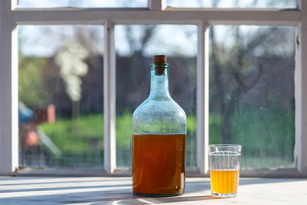 एक गर्म वसंत के दिन खिड़की पर किण्वित बिर्च सैप से बने पेय के साथ बड़ी बोतल, बंद करें। पारंपरिक यूक्रेनी शीत जौ यार्ड के पास टेबल पर एक ग्लास जार और ग्लास में क्वास पीते हैं स्टॉक फ़ोटो
