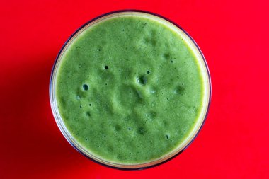 Kırmızı arka planda bir bardak yeşil smoothie, yakın plan, üst manzara. Muz, dereotu, maydanoz ve avokadodan yapılan sağlıklı organik içecek. Yeşil bir içeceğin yüzeyinde köpükler ve kabarcıklar.