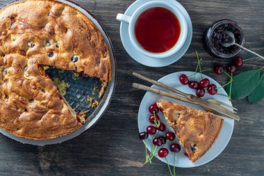 Bir parça vişneli turta, yanında taze kirazlar ve ahşap bir masada beyaz bir fincan çay, üst manzara, yakın çekim. Lezzetli ev yapımı kirazlı Amerikan pastası. Kahvaltıda tatlı