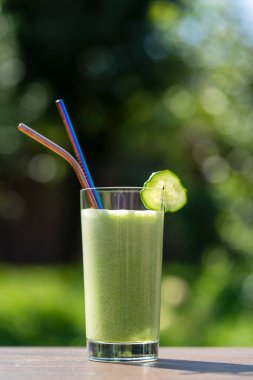 Doğanın arka planındaki kafenin ahşap masasında bir bardak yeşil smoothie, yakın plan. Muz, dereotu, maydanoz, salatalık ve avokadodan yapılan sağlıklı organik içecek.