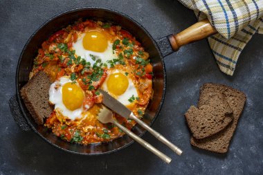 Shakshuka in frying pan for breakfast, close up. Homemade shakshuka from fried eggs, onion, bell pepper, red tomato and parsley in a cast iron skillet, top view clipart