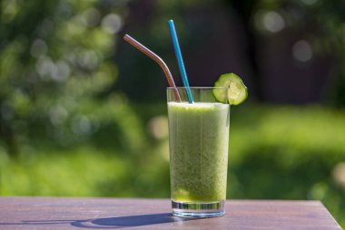 Doğanın arka planındaki kafenin ahşap masasında bir bardak yeşil smoothie, yakın plan. Muz, dereotu, maydanoz, salatalık ve avokadodan yapılan sağlıklı organik içecek.