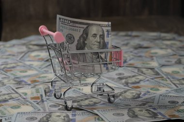 Tekerlekli alışveriş arabası ve ahşap arka planda bir sürü 100 dolarlık banknot. Finans, iş, alım-satım, harcama ve satış kavramı