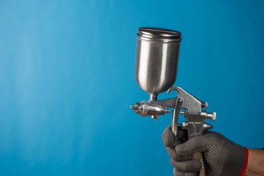 Boyacı, endüstriyel boyama ve kaplama için kullanılan endüstriyel boyutta havasız sprey tabancayı eldiven içinde tutuyor. Gümüş bardaklı sprey tabanca ve araba parçaları ya da mavi arka planda mobilya boyamak için sapı
