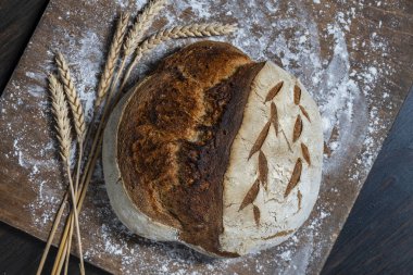 Tahta kesme tahtasının üzerinde buğday kulaklı taze ekşi ekmeğin yuvarlak somunu, yakın çekim, üst manzara. Rustik ekşi ekmek. Evde sağlıklı ekşi ekmek pişirmek.
