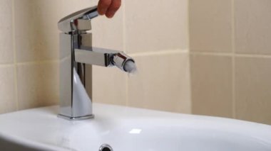 Modern banyodaki bej kaplamalı musluktan akan su ile birlikte beyaz seramik bidet detayları. Basit stil ve modern ev dekorasyonu. Kapat.