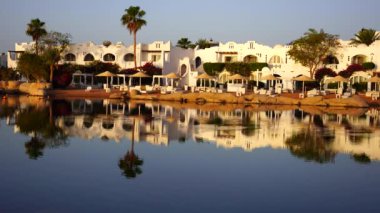 Gündoğumunda, Mısır 'ın Sharm El Sheikh tatil beldesindeki sakin deniz suyuna binalar, güneşlikler ve şemsiyeler yansıtılır.