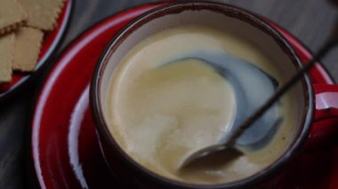 Sabaha karşı, ahşap masa arkasında kırmızı bardakta kaşık ile karıştırılmış sıcak kahve köpüğü, kapat.