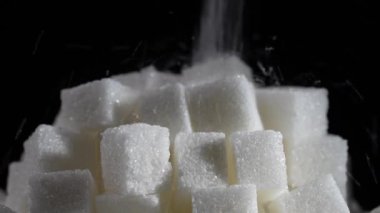 Beyaz granüllü şeker siyah arkaplandaki şeker küplerinin üzerine düşer. Ağır çekim. Sağlıksız yeme, obezite, kalp hastalığı, diyabet kavramı.