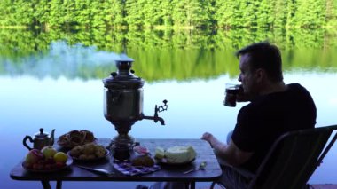Gölün kenarında çay içen bir adam. Sabah Ukrayna 'da yeşil ormandaki sakin su gölünün yanındaki masada beyaz duman ve yiyecek bulunan klasik metal çay semaveri.