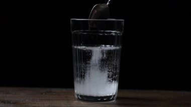Beyaz şekeri kaşıktan bardağa döküyorum. Siyah arka plandaki ahşap masada su var. Yaklaş, yavaş çekim.