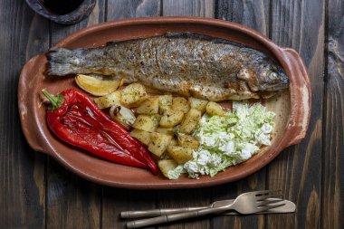 Ahşap masadaki kil tabakta kırmızı biberli, patatesli ve Çin lahana salatalı lezzetli alabalık balığı.