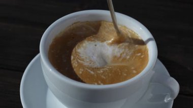 Tahta masada beyaz bardakta kaşıkla karıştırılmış koyu köpüklü sıcak kahve kapuçinosu, yakın çekim. Süt köpüğü kapuçino, latte.
