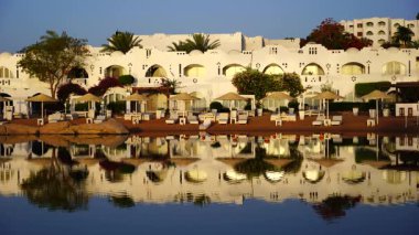 Gündoğumunda, Mısır 'ın Sharm El Sheikh tatil beldesindeki sakin deniz suyuna binalar, güneşlikler ve şemsiyeler yansıtılır.