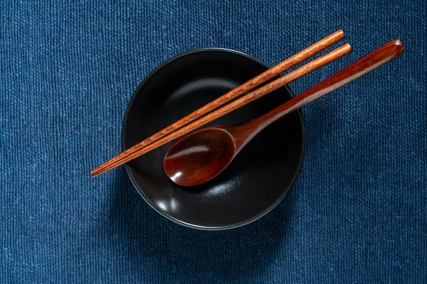 Bir çift ahşap yemek çubuğu ve kaşık mavi kumaş arka planda boş bir çorba kasesinde yatıyor.