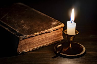 Geceleri karanlık odada, eski açık kitabın yanında, ahşap masanın üzerinde bronz şamdanda yanan mum, kapat. Eski kitapların yanında mum yakan klasik bir hayat.