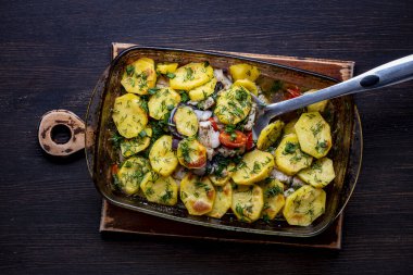 Dilimlenmiş patates, balık fileto, domates, soğan ve serpilmiş otlar ahşap bir masada cam bir fırın kabında, yakın çekim, üst görüş