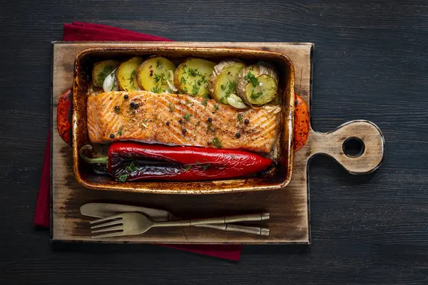 Kırmızı biberli fırında somon filetosu ve seramik fırın kabında patates dilimleri ahşap masada tahta kesme tahtasında, yakın çekim, üst manzara. Yemekte sebzeli balık var.