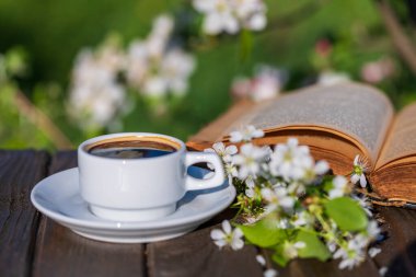 Beyaz bir fincan sıcak kahve, beyaz kiraz çiçekleri ve bahar bahçesi arka planına karşı eski bir kitapla ahşap masanın üzerinde. Kapat. Yumuşak romantik bir ortam, bahar mevsiminde kahve partisi.