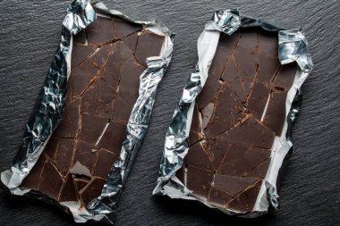 Siyah arka planda folyoyla kaplanmış iki kırık çikolata, yakın plan, üst manzara. Tatlı yemek.