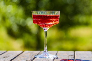 Kaliteli kristal bardakta parlak kırmızı bir kokteyl. Yaz bahçesi arka planında, ahşap masada. Yaklaş. Güneşli bir günde rahatlamak için nefis bir alkollü içecek.