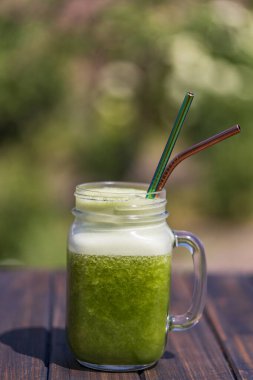 Cam bardakta taze yeşil meyve püresi. Yazlık bahçe arka planında metal kamışla ahşap masada, yakın plan. Sağlıklı beslenme kavramı. Yapraklı yeşillerden, otlardan, sebzelerden ve meyvelerden arınma içeceği