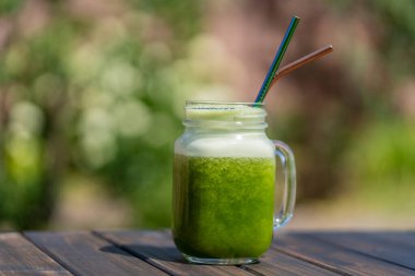 Cam bardakta taze yeşil meyve püresi. Yazlık bahçe arka planında metal kamışla ahşap masada, yakın plan. Sağlıklı beslenme kavramı. Yapraklı yeşillerden, otlardan, sebzelerden ve meyvelerden arınma içeceği